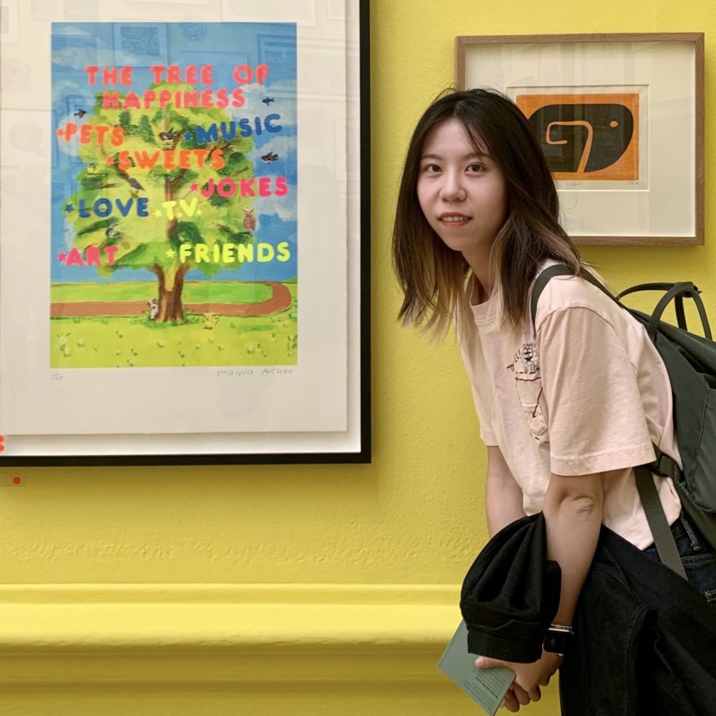 Yixin has shoulder length dark hair and is standing in side profile wearing a pink t-shirt. She is in a museum next to an artwork with the text: The tree of happiness, Pets, Music, Sweets, Love, Jokes, TV, Art, Friends atop a painted tree with a winding pathway.
