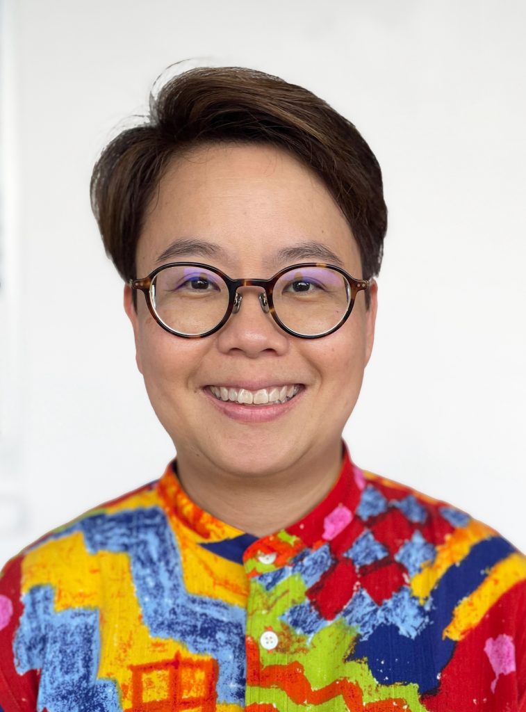 Hui May has short dark hair and round black glasses. She is wearing a colourful shirt.